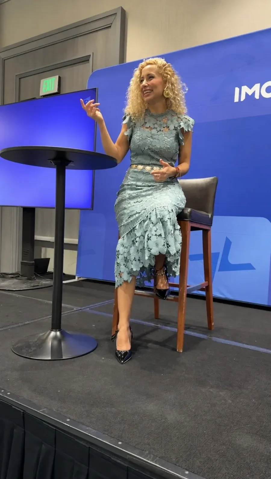 Leslye Rendón compartiendo una intervención, con micrófono de diadema, ante el fondo de un encuentro IMC de Maxwell Leadership.